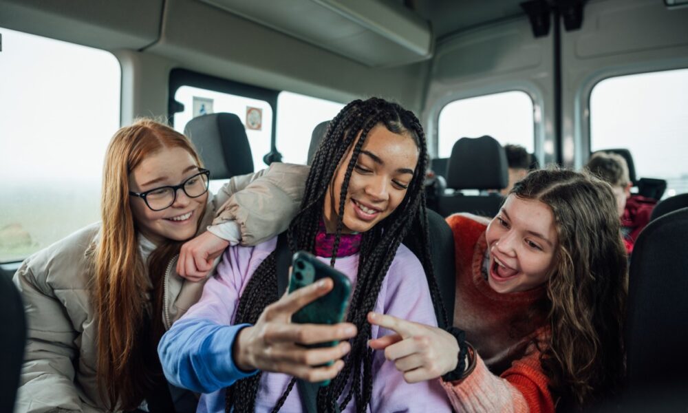 A front view of a group of three teenage girls on the minibus on the way to go on a hiking field trip. They are looking at one of the girls mobile phones and smiling and pointing at the screen.