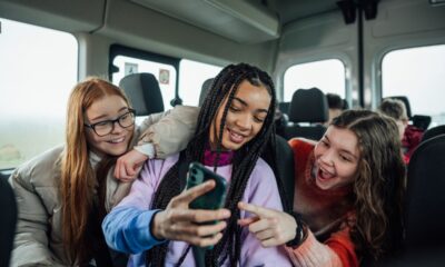 A front view of a group of three teenage girls on the minibus on the way to go on a hiking field trip. They are looking at one of the girls mobile phones and smiling and pointing at the screen.