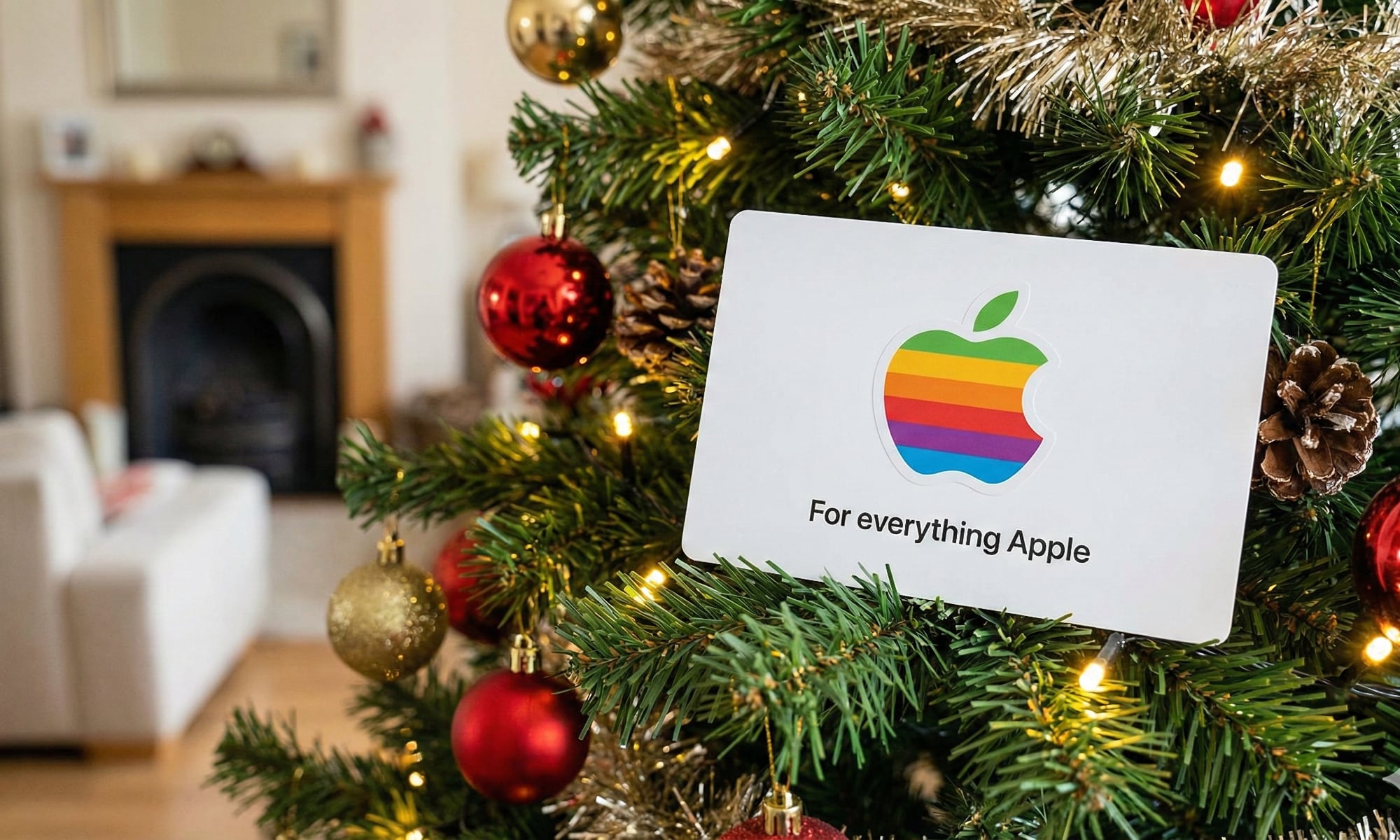 A modern, unified Apple Gift Card with the colorful rainbow logo and "For everything Apple" text, resting on the branches of a decorated Christmas tree amidst warm lights, red baubles, and pinecones.