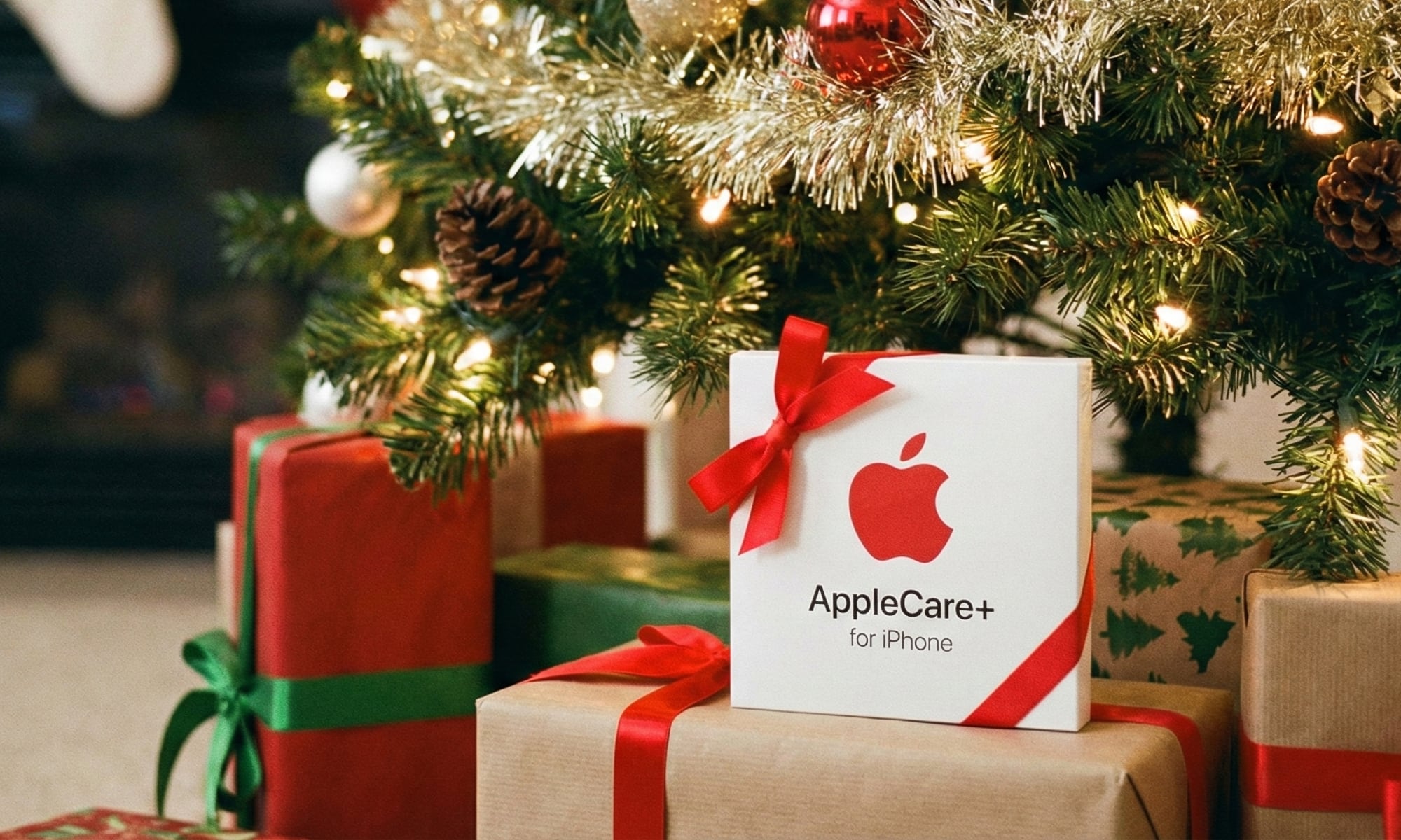 A white AppleCare+ package tied with a red ribbon sits prominently among other wrapped gifts under a brightly lit and decorated Christmas tree, with stockings hanging on a fireplace in the background.