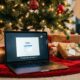 An open MacBook Pro displaying the "Hello" setup screen, sitting on a red tree skirt under a decorated Christmas tree with wrapped gifts.