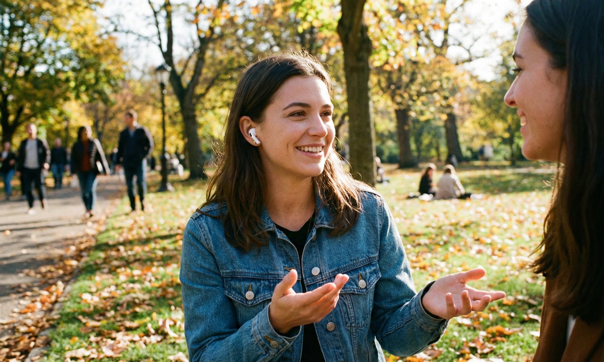 A smiling woman in a denim jacket wearing AirPods Pro earbuds and gesturing while having a conversation with a friend in a sunny autumn park, demonstrating the Conversation Awareness feature.