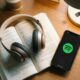 An iPhone showing a Spotify interface next to an open paperback book with over-ear headphones resting on the pages.