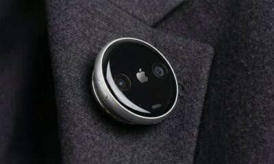 A concept design of a circular Apple AI pin attached to a charcoal grey suit lapel. The device features a polished aluminum rim, a black glass face with a small silver Apple logo in the center, and two visible camera lenses on either side. A physical button is visible on the side of the thin, disc-shaped wearable.