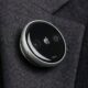 A concept design of a circular Apple AI pin attached to a charcoal grey suit lapel. The device features a polished aluminum rim, a black glass face with a small silver Apple logo in the center, and two visible camera lenses on either side. A physical button is visible on the side of the thin, disc-shaped wearable.