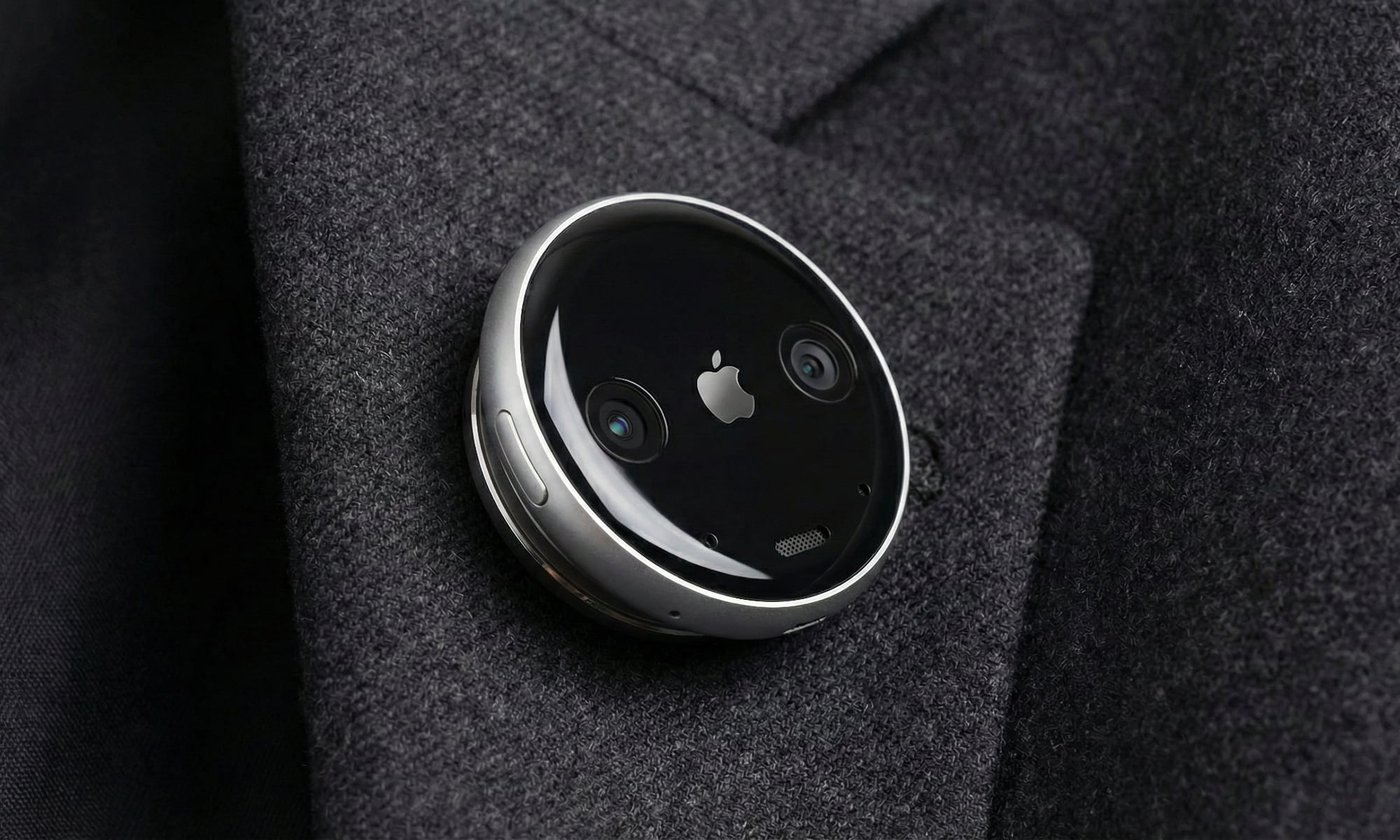A concept design of a circular Apple AI pin attached to a charcoal grey suit lapel. The device features a polished aluminum rim, a black glass face with a small silver Apple logo in the center, and two visible camera lenses on either side. A physical button is visible on the side of the thin, disc-shaped wearable.