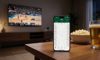 An iPhone 17 Pro on a wooden coffee table displaying the Apple Sports app showing NCAA tournament brackets, with a basketball game on a TV in the blurred background.