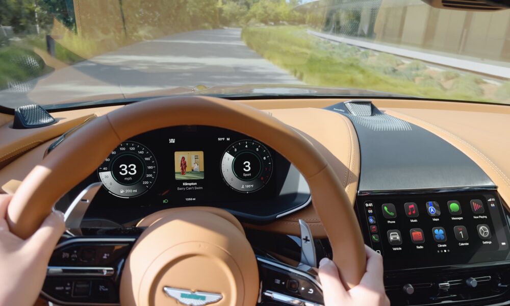 A professional press photo of the Apple CarPlay Ultra interface inside an Aston Martin. The dashboard features two distinct digital displays: a 10.25-inch central touchscreen showing a grid of icons including Phone, Music, Messages, and new vehicle-integrated apps for Radio and Climate. Behind the steering wheel, the driver’s instrument cluster is fully overtaken by CarPlay, displaying traditional-style Aston Martin circular dials for speed and RPM with a live Apple Maps navigation view in the center. The cabin features premium silver switchgear and leather-stitched trim.
