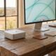 A silver Apple Mac mini sits on a rustic wooden desk in a sunlit home office with a large window overlooking a Texas landscape. A monitor, keyboard, mouse, and a leather coaster with a star emblem are also on the desk.