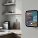 A concept image of a sleek, square Apple smart display (HomePad) magnetically attached to a modern kitchen wall, displaying a personalized dashboard with Siri, calendar appointments, weather, and home controls.