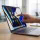 A close-up lifestyle concept shot of a person’s hand using touch gestures on the ultra-slim OLED display of a 2026 MacBook Pro in a bright, modern office setting.