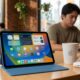 Concept image of a 12th-generation iPad in blue sitting on a wooden coffee shop table next to a latte and a pair of AirPods, showing the Liquid Glass interface of iPadOS 26.