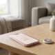 A soft pink iPhone 17e sitting on a wooden coffee table next to a 2nd-gen AirTag, with a blurred background of a modern living room.