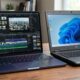 A side-by-side comparison shows the incredibly thin Indigo MacBook Neo editing 4K video in Final Cut Pro next to a significantly thicker, bulky black textured plastic budget PC laptop, highlighting the design and performance gap.