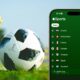 iPhone Showing Apple Sports app with FIFA 2026 World Cup Countries against a FIFA trophy and ball sitting on a field.