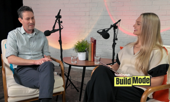 A podcast-style interview setup shows a man and a woman seated across from each other in armchairs, speaking into microphones. They sit beside a small round table with water glasses, books, and a potted plant, against a white brick wall backdrop. A “TechCrunch Build Mode” logo appears in the lower corner.