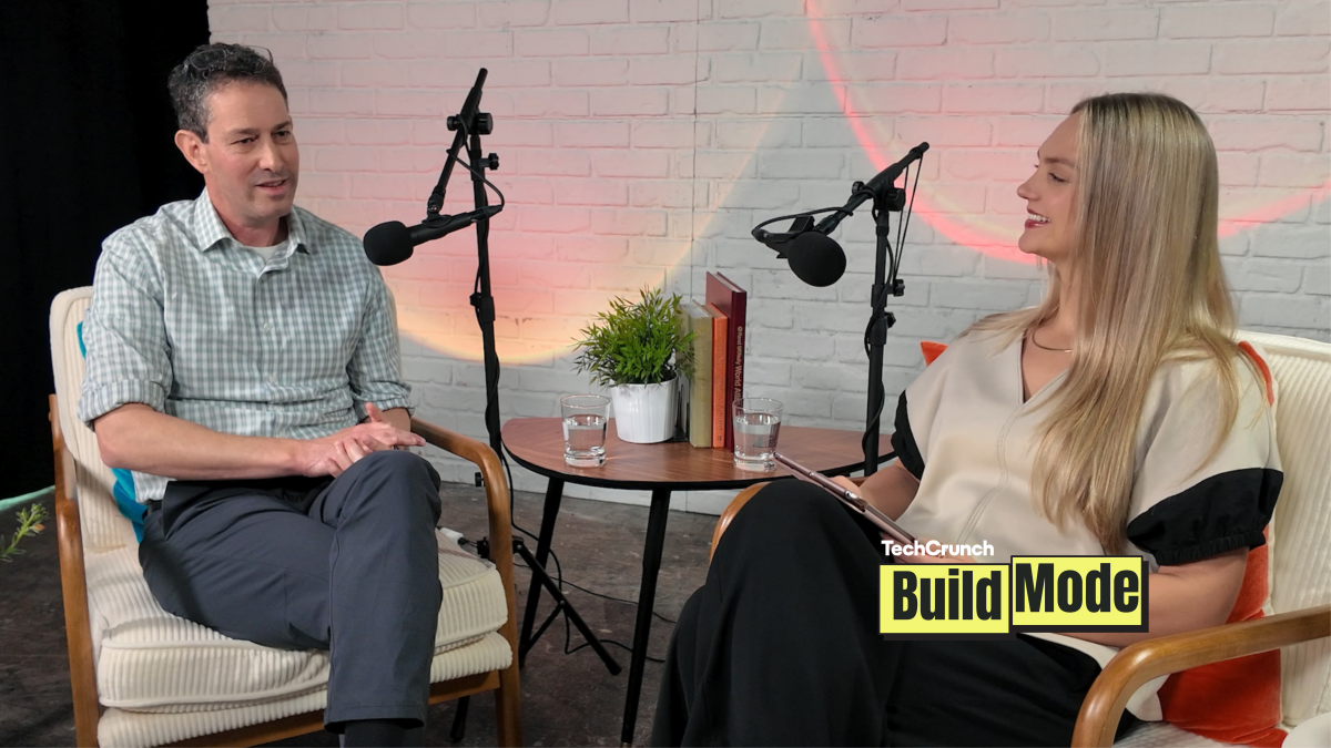 A podcast-style interview setup shows a man and a woman seated across from each other in armchairs, speaking into microphones. They sit beside a small round table with water glasses, books, and a potted plant, against a white brick wall backdrop. A “TechCrunch Build Mode” logo appears in the lower corner.