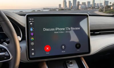 A photorealistic interior of a modern car showing a large dashboard infotainment screen running Apple CarPlay. The display features the Google Meet app interface with an active call titled "Discuss iPhone 17e Review." The background through the windshield shows a scenic coastal road and a city skyline at sunset.
