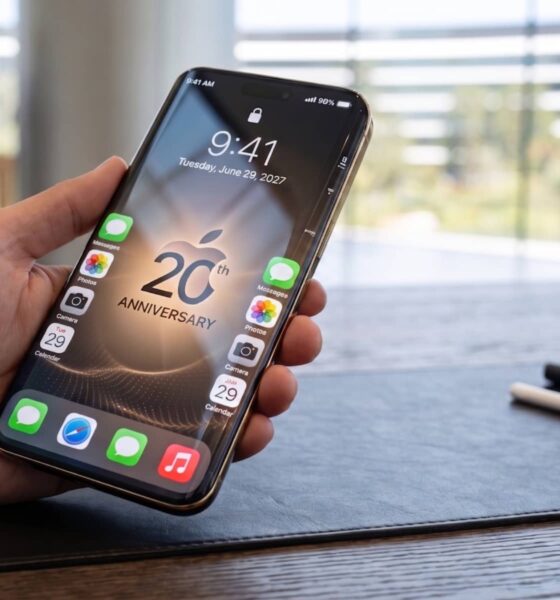 A detailed, realistic landscape photo of a hand holding the modern, sleek 'iPhone 20' concept. The phone screen displays a 2027 date and a special '20th Anniversary' wallpaper. The surrounding desk includes a Steve Jobs portrait and Apple Pencil on a seamless leather mat.