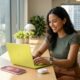 A diverse young professional smiling while working at a modern, sunlit desk with the 'Citrus' MacBook Neo, soft pink iPhone 17e, Apple Watch SE 3, and AirPods 4 arranged together, showcasing a complete and affordable Apple ecosystem in 2026.