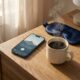 An iPhone resting on a natural wood nightstand next to a steaming artisanal coffee mug and a dark navy blue silk sleep mask. The phone screen displays a calming 2026 meditation app with text 'START YOUR DAY CALM' and '7:30 AM 15 min session'. The background shows softly blurred bedroom linens and warm morning window light.