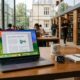 A MacBook Neo and an Apple Watch on a university library desk with a blurred backgroudn of students studying in a modern library setting.