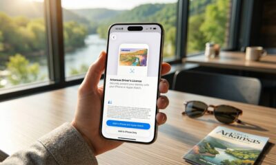 A close-up lifestyle shot of a person holding an iPhone that displays the Arkansas driver's license enrollment screen for Apple Wallet, set against a wooden cafe table with sunglasses and a scenic river window view.