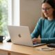 A woman working on a silver MacBook at a bright wooden desk, with a wireless keyboard, mouse, and an unplugged USB-C charging cable nearby, illustrating portable laptop battery usage.