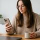 A young woman calmly focuses on her customized 'Work' Lock Screen on an iPhone 17 Pro, sitting at a clean light oak desk in a bright home office with a coffee mug, MacBook, and notebook. The background is a soft bokeh.
