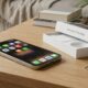 A lifestyle photograph of a newly unboxed iPhone 17 Pro Max on a wooden living room coffee table. The phone shows rows of app icons like Spotify, Signal, and 1Password against a warm living room background.
