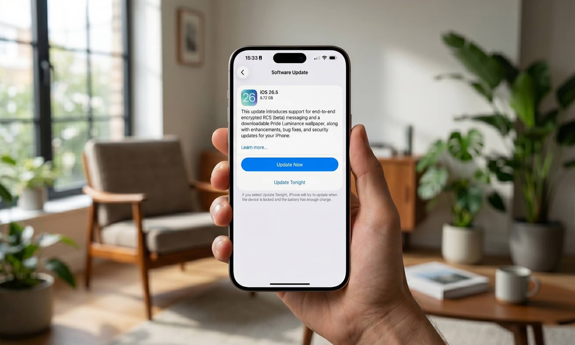 A close-up photograph of a person in a modern, plant-filled living room, holding an iPhone showing the iOS 26.5 Software Update screen. The screen clearly details the new features, including end-to-end encrypted RCS messaging (beta).