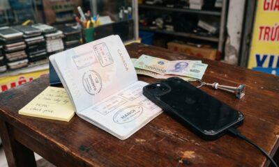 A black iPhone plugged into a charging cable on a rustic wooden desk inside a bustling Vietnamese electronics repair shop. Next to the device lies an open Canadian passport displaying travel stamps from Toronto and Hanoi, along with Vietnamese Dong banknotes and a yellow sticky note reading "HCMC - check price".