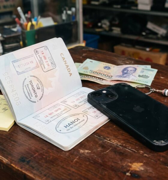 A black iPhone plugged into a charging cable on a rustic wooden desk inside a bustling Vietnamese electronics repair shop. Next to the device lies an open Canadian passport displaying travel stamps from Toronto and Hanoi, along with Vietnamese Dong banknotes and a yellow sticky note reading "HCMC - check price".