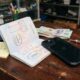 A black iPhone plugged into a charging cable on a rustic wooden desk inside a bustling Vietnamese electronics repair shop. Next to the device lies an open Canadian passport displaying travel stamps from Toronto and Hanoi, along with Vietnamese Dong banknotes and a yellow sticky note reading "HCMC - check price".