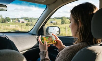 An over-the-shoulder photograph of a passenger in a car holding an iPhone and playing Stardew Valley, with a scenic, sunny country road and farm fields visible through the vehicle’s windows.