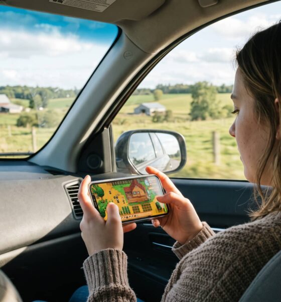 An over-the-shoulder photograph of a passenger in a car holding an iPhone and playing Stardew Valley, with a scenic, sunny country road and farm fields visible through the vehicle’s windows.