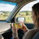 An over-the-shoulder photograph of a passenger in a car holding an iPhone and playing Stardew Valley, with a scenic, sunny country road and farm fields visible through the vehicle’s windows.