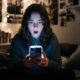 A teenager sitting on a bed in a darkened bedroom, looking down at an iPhone screen with a shocked expression, illustrating the real-world risks of unmoderated explicit content on minor-registered devices.