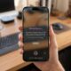 A first-person close-up of a hand holding an iPhone 17 Max in a bright, modern home office with a blurred wooden desk background. The iPhone screen displays the new iOS 27 "Siri in Camera" interface, showcasing the native Visual Intelligence onboarding text and the dedicated Siri camera shooting mode.