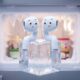Whimsical cartoon animation visualization of a pair of white wireless earbuds, in the style of AirPods with large expressive eyes, perched on top of a single, clear, frosted ice cube inside a very cold domestic freezer compartment. The background is a frosted freezer interior.