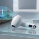 A high-end conceptual photograph of two white wireless earbuds resembling Apple AirPods resting on a reflective glass surface. The foreground earbud clearly displays a dual-camera lens array and an active amber indicator light on its stem. A glowing blue holographic interface projects an 'i' icon and scanning rings over a stack of old books, one of which is titled 'APPLE'S VISUAL INTELLIGENCE: RUMOR TO REALITY.' Data code reflections are visible on the surface.