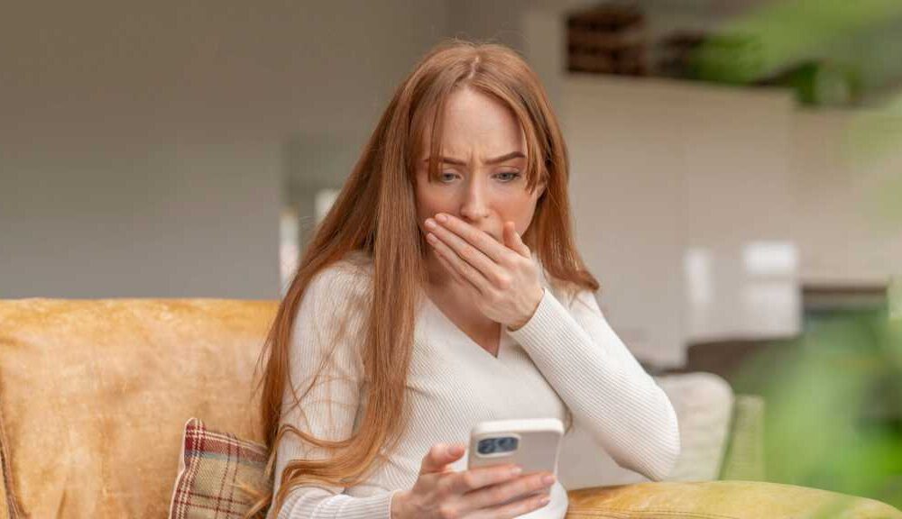 Rothaarige junge Frau blickt ängstlich mit Hand vor dem Mund auf ihr Handy, während sie auf dem Sofa sitzt.