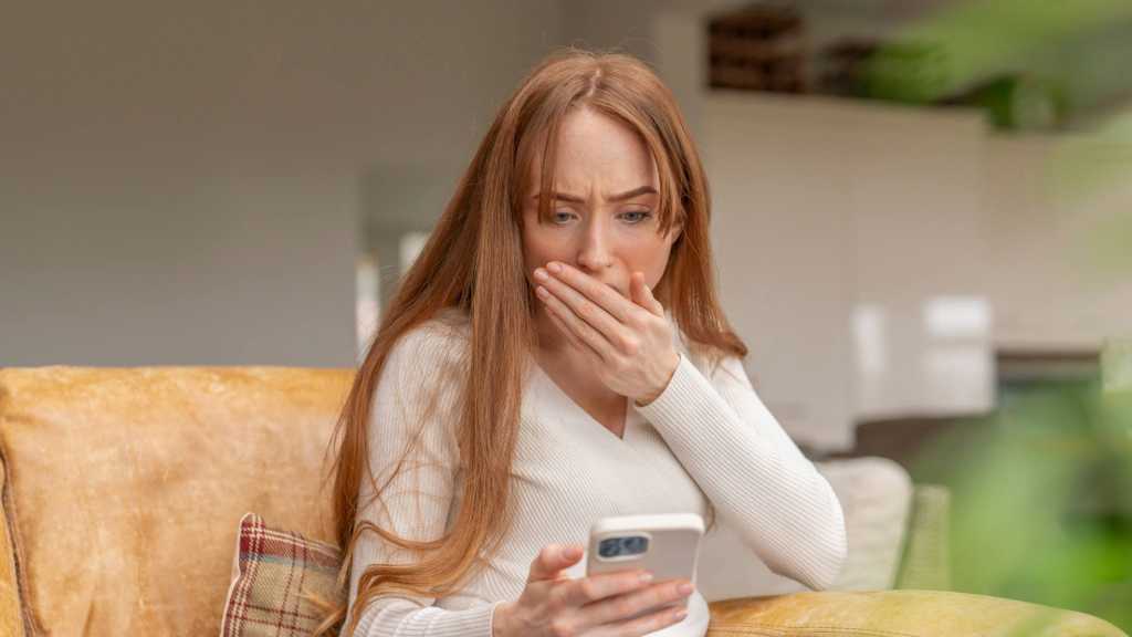 Rothaarige junge Frau blickt ängstlich mit Hand vor dem Mund auf ihr Handy, während sie auf dem Sofa sitzt.