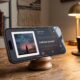 A photo of an iPhone Pro propped on a wooden stand in a cozy home office, displaying the new Apple Music landscape "Now Playing" screen feature coming in iOS 27. The screen shows album art and lyrics for "Fear No More" by Building 429. A coffee mug and lamp are in the background.