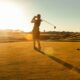 A silhouette of a person holding a swung golf club while teeing off into the sunset.