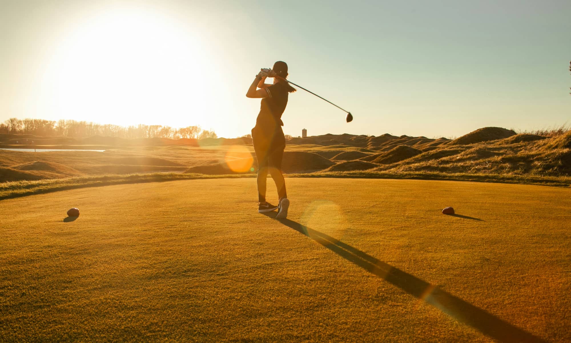 A silhouette of a person holding a swung golf club while teeing off into the sunset.