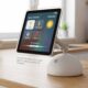 A detailed concept rendering of Apple's rumored J490 smart home hub, featuring a 6-inch square display mounted on an articulated arm that sits on a white, half-dome speaker base, reminiscent of the 2002 iMac G4 design, resting on a clean oak wood countertop.