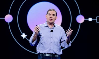 Matt Garman speaks on stage in a light purple button-up shirt, gesturing with both hands and holding a presentation clicker, in front of a dark screen showing a purple circle with orbit lines.