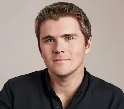 Headshot of Stripe co-founder and President John Collison, wearing a dark collared shirt against a light gray background.