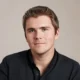 Headshot of Stripe co-founder and President John Collison, wearing a dark collared shirt against a light gray background.